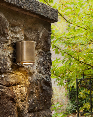Bronze outdoor wall light fixture on a stone wall with greenery in the background