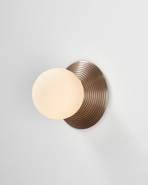 Wall-mounted light fixture with a round, ribbed bronze base and a white glass shade on a light gray background.