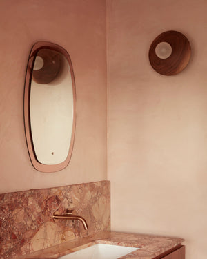 Bathroom corner with a mirror, faucet, and sink against a pink wall and a wooden wall sconce