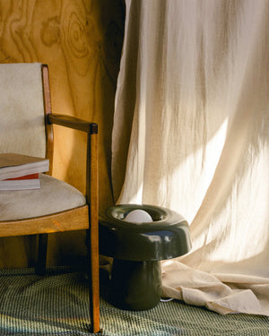 Wooden chair with a white cushion next to a greenlamp on a textured surface with a curtain in the background.