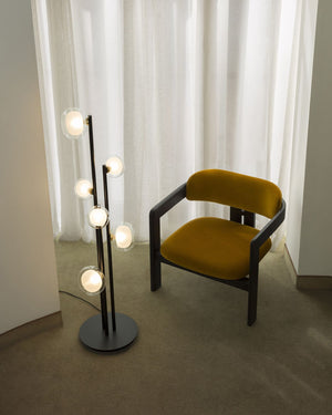Modern chair with mustard-colored seat and backrest next to a floor lamp with multiple lights in a room with white curtains.