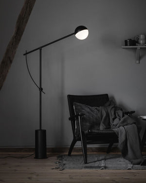 Pivoting Steel Matte Black Floor Lamp with a light on in a dimly lit room with a chair and blanket.