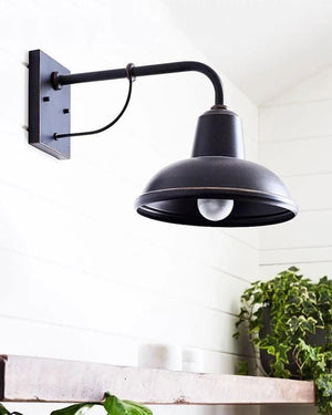 Black wall-mounted barn-style light fixture on a white wall with plants below.