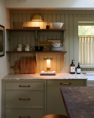 Brass shell wall light mounted above open kitchen shelf casting warm downlight over marble benchtop in sage green kitchen