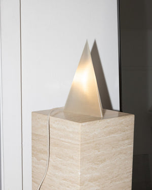 A modern sculptural table lamp featuring a glowing, translucent triangular prism on top of a textured beige stone or marble pedestal, shown against a neutral indoor background.
