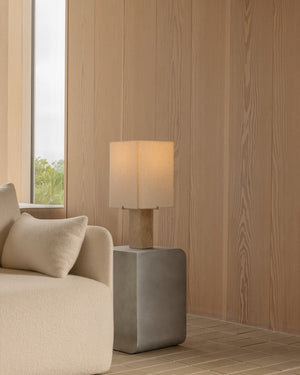 Medium narrow travertine base with fabric shade with warm glowing light on a table in a minimal living room interior