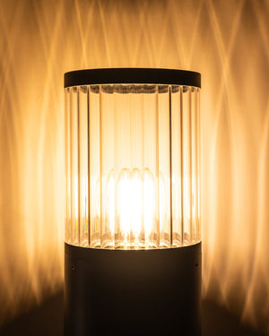 The bollard lit after dark, throwing bands of warm light up a textured wall behind it.