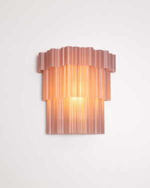 A modern wall sconce light fixture with a translucent, ribbed, tiered design in a dusty rose pink color, emitting a warm orange glow against a plain white wall.