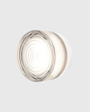 A modern circular wall light fixture with a clear glass shade and a white inner diffuser, emitting a warm glow against a light gray background.