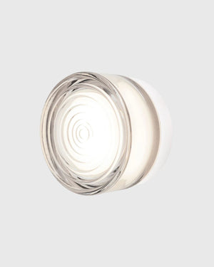 A modern circular wall light fixture with a clear glass shade and a white inner diffuser, emitting a warm glow against a light gray background.
