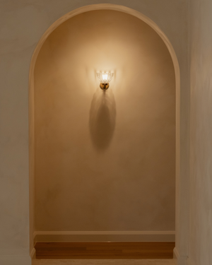 A warm floral wall sconce with a gold-toned brass fixture and clear glass shade, mounted on a beige plaster wall inside an arch.
