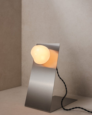 Lean Table Lamp with its cantilevered steel stand upright and glass bulb unlit, on a studio backdrop