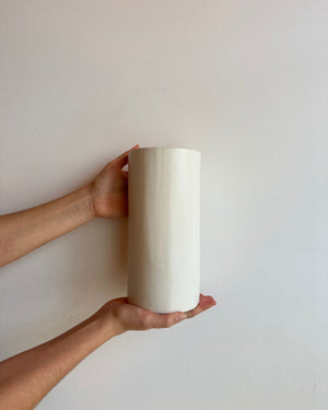 A person's hands holding a plain white cylindrical ceramic wall light against a neutral off-white wall.