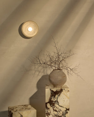 Decorative vase with branches on a marble pedestal against a textured wall with a light fixture.