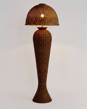 An hourglass rattan floor lamp with a woven dome shade and matching tapered base, shown lit against a plain light background.