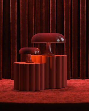Two mushroom-shaped table lamps in a glossy deep red finish, displayed on matching fluted pedestal tables against a dark red velvet curtain backdrop.