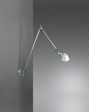 A modern wall-mounted adjustable arm lamp with a silver metal articulated arm and conical shade, shown against a dark gray wall with light casting on the right.