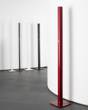 A modern minimalist floor lamp with a tall cylindrical red column and a square base, standing in a minimalist room with gray tiled floors and white walls. Four other floor lamps in black and white are visible in the background.