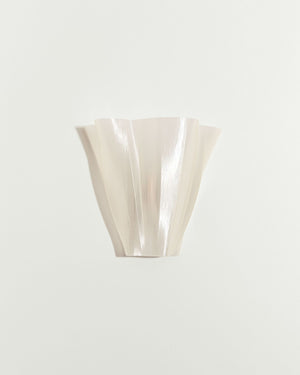 A sculptural, fluted wall sconce in a pearlescent off-white finish, mounted on a plain light gray wall.