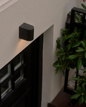 Black outdoor wall light fixture on a building exterior with plants in the background.