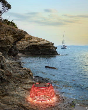 Red woven lantern on a rocky cliff by the ocean with a sailboat in the background.