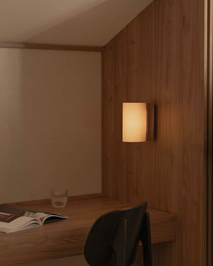 Curved papyrus shade walnut wall light in short size with warm glowing light mounted on a wooden panel next to a desk