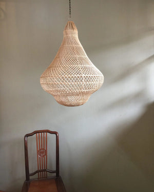 Woven pendant light hanging against a plain wall with a wooden chair in the foreground.