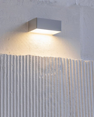 A modern rectangular wall sconce light fixture in a matte gray finish, mounted on a textured white plaster wall and casting a warm yellow light downward.
