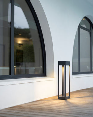 A modern black rectangular outdoor bollard with a warm glowing light inside, positioned on a wooden deck next to a white building with arched windows.