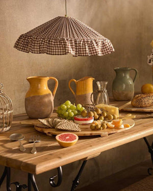 A rustic wooden dining table with cheeseboard, pitchers ad bread styled with a ruffle trim pleated pendant in brown gingham fabric 