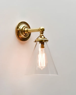 A wall-mounted brass sconce with a clear glass cone shade and a glowing filament bulb, shown against a plain light wall.