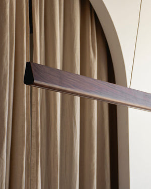 Close-up of walnut timber linear LED pendant end cap detail suspended in arched interior with draped fabric backdrop