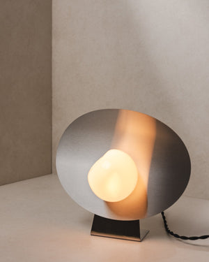 Wink Table Lamp with its steel oval upright on a slim stand, opal glass bulb lit